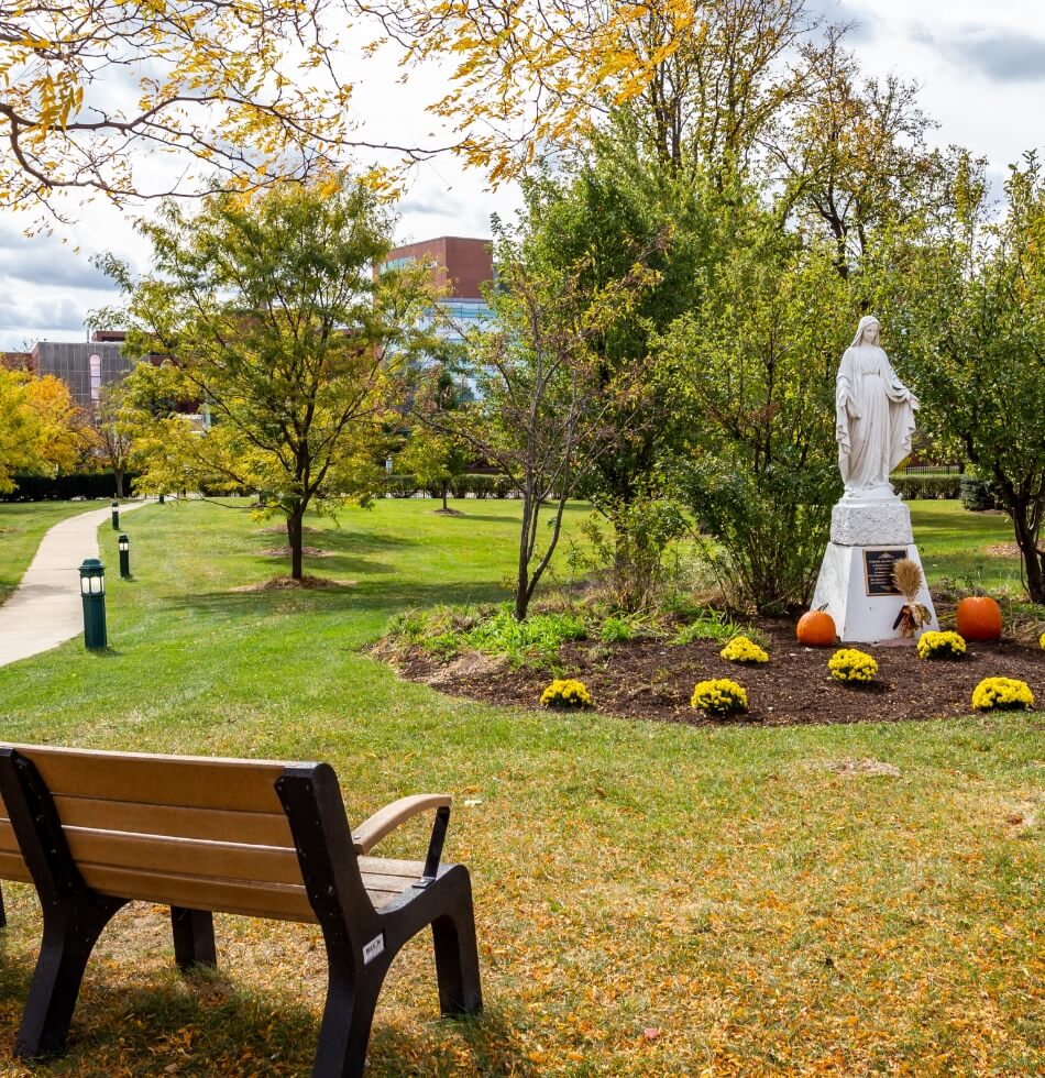 Peaceful garden scene with Virgin Mary statue, bench, and fall foliage at Amazing Grace Senior Living