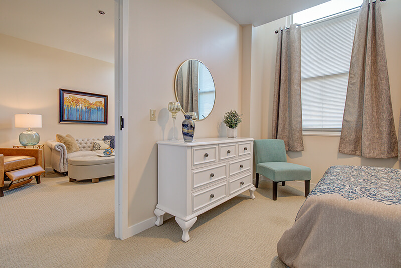One-bedroom apartment interior showing dresser, accent chair, and natural light at Amazing Grace Senior Living