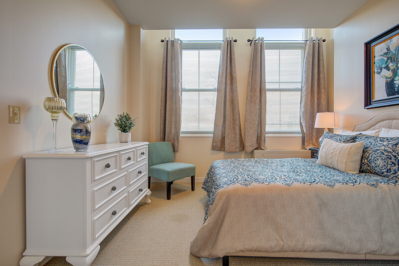 Bright one-bedroom apartment with large windows, decorative bedding, and modern furnishings at Amazing Grace Senior Living