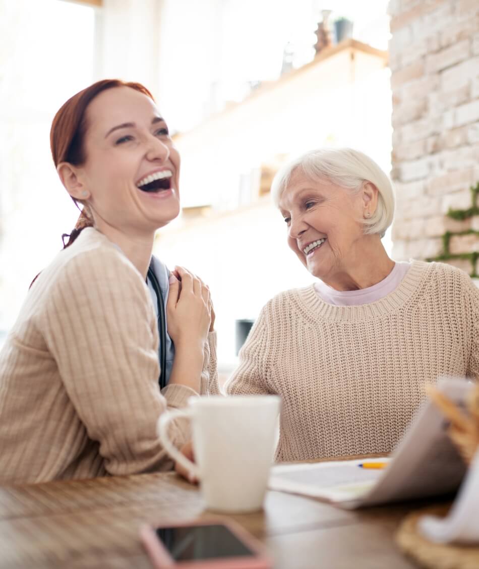 Smiling senior woman enjoying a cheerful conversation with her caregiver over coffee at Amazing Grace Senior Living