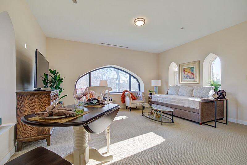 Bright studio apartment with large arched window, dining space, and comfortable seating at Amazing Grace Senior Living