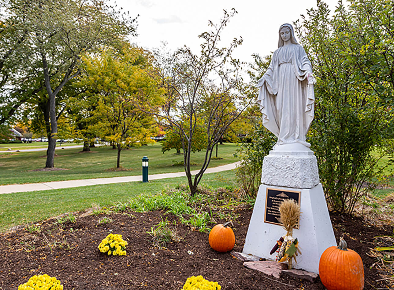 Virgin Mary statue surrounded by fall decorations and flowers in the garden at Amazing Grace Senior Living