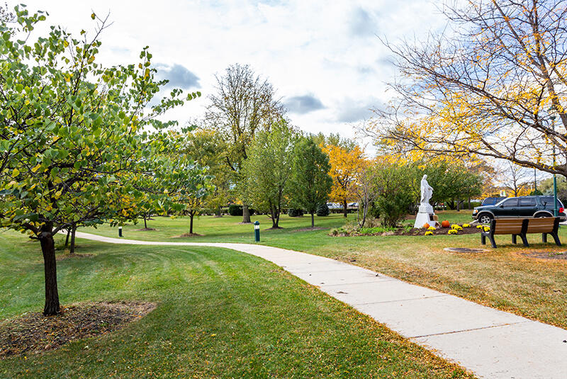 Peaceful landscaped walking path with trees and statue on the grounds of Amazing Grace Senior Living